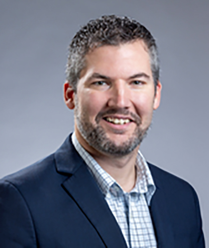 A man with short gray hair and a beard is wearing a dark suit jacket over a checkered shirt, embodying the confident charm of a transactional advisory expert. He smiles warmly at the camera against a plain gray background.