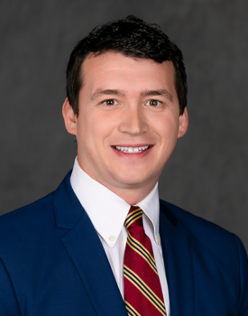 Scott in a blue suit, white shirt, and striped red tie smiles in front of a gray backdrop, exuding the confident poise of a transactional advisory expert.