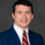 Scott in a blue suit, white shirt, and striped red tie smiles in front of a gray backdrop, exuding the confident poise of a transactional advisory expert.