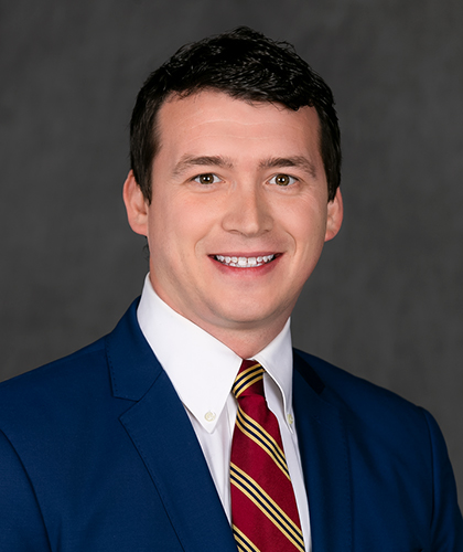 Scott in a blue suit, white shirt, and striped red tie smiles in front of a gray backdrop, exuding the confident poise of a transactional advisory expert.