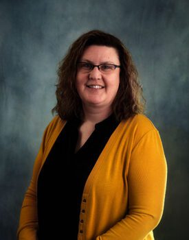 A person with shoulder-length curly hair, wearing glasses, a black top, and a mustard yellow cardigan, smiles confidently as if ready for a transactional advisory session against the dark, mottled background.