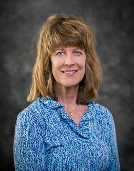 A woman with shoulder-length hair, wearing a blue patterned blouse, is smiling confidently against a gray textured background, embodying the spirit of transactional advisory expertise.