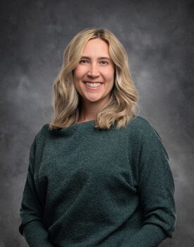 A person with shoulder-length blonde hair, exuding confidence akin to a transactional advisory expert, is smiling warmly in a green sweater. The backdrop is a moody, cloudy gray sky.