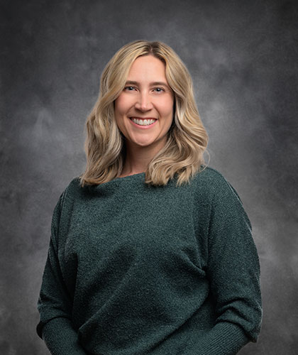 A person with shoulder-length blonde hair, exuding confidence akin to a transactional advisory expert, is smiling warmly in a green sweater. The backdrop is a moody, cloudy gray sky.