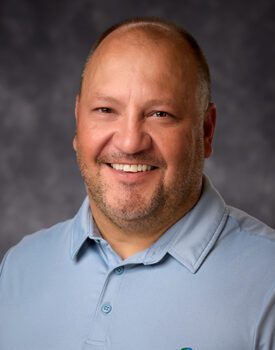Smiling man in a light blue polo shirt, exuding confidence and expertise in transactional advisory, against a gray background.
