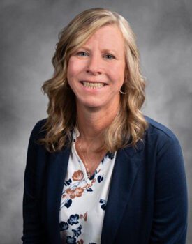 Smiling woman with blonde, wavy hair, wearing a navy blazer over a floral blouse, exuding professionalism and confidence, suggesting expertise in transactional advisory against a gray background.