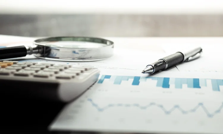 Forensic_Accounting_magnifying_glass_750x450 Close-up of a calculator, magnifying glass, pen, and financial chart with blue bars and a line graph on a desk, tools essential for forensic accounting analysis.