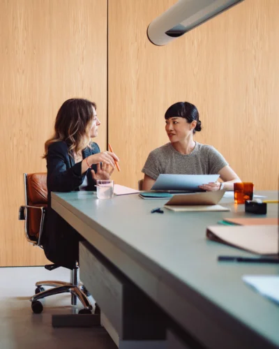 Two women sit at a long table in a modern office setting, deeply engaged in discussion. One holds a pen and notepad, while the other examines data on her tablet—both immersed in the meticulous world of forensic accounting.