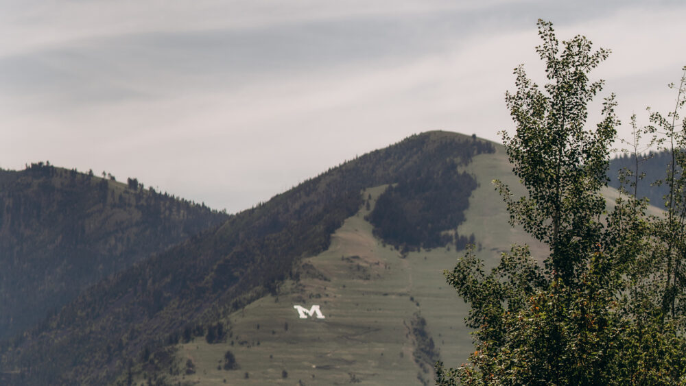 Mountain with a large white "M" on its slope, surrounded by trees and a cloudy sky.