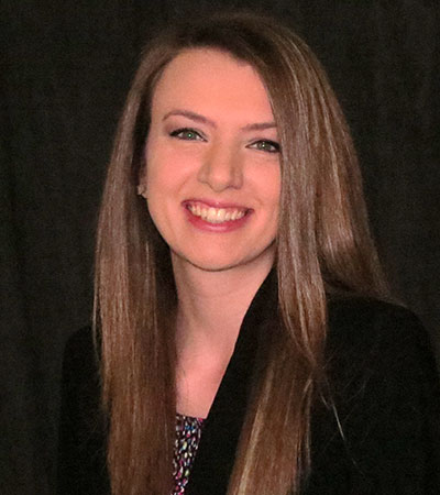 A person with long brown hair and wearing a black blazer smiles against a dark background.
