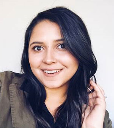 A person with long dark hair smiles at the camera, wearing a dark top and olive jacket, with a light background.