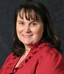 Roxie Azparren, a woman with shoulder-length dark hair, is wearing a red ruffled blouse and smiling against a dark backdrop.
