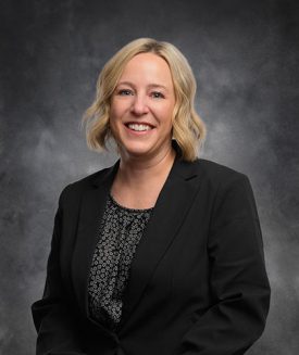 Brea Bauer, with short blonde hair, smiles warmly as she dons a black blazer over a patterned blouse. She stands elegantly against a dark, neutral backdrop.