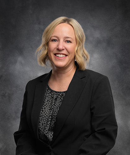 Brea Bauer, with short blonde hair, smiles warmly as she dons a black blazer over a patterned blouse. She stands elegantly against a dark, neutral backdrop.