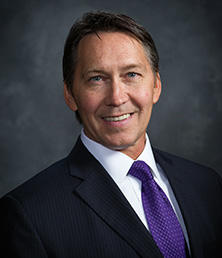 Kevin Bearley, a man in a dark suit with a white shirt and purple tie, smiles confidently against a dark gray background.