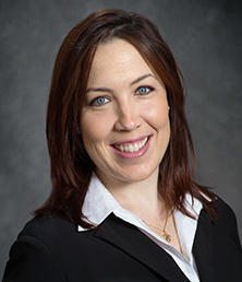 Rachel Biggs, with shoulder-length brown hair, is wearing a black blazer and white shirt and smiling against a gray background.