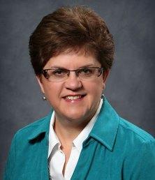 Person with short brown hair and glasses, resembling Jeannie Blue, wearing a teal jacket and white shirt, stands against a gray background.