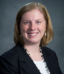 Stephanie Bolte-Lank, with her short light brown hair and radiant smile, exudes confidence in a black blazer, white top, and pearl earrings against a muted gray backdrop.