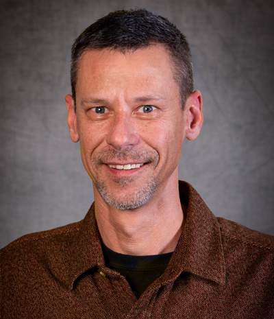 Brian Holder, with his short hair and trimmed beard, smiles warmly in a brown collared shirt against a plain gray background.