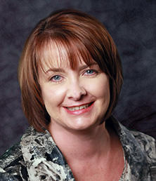 Dawn Buchanan, with her short brown hair and patterned top, smiles warmly against a dark textured backdrop.