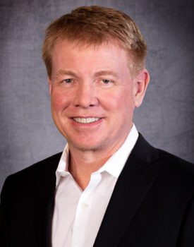 Chris Haverkamp, with short light brown hair, wearing a black suit and white shirt, smiles warmly against a gray background.