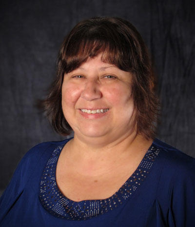 Colleen Parsons, a smiling woman with shoulder-length brown hair, is wearing a blue blouse against a dark background.