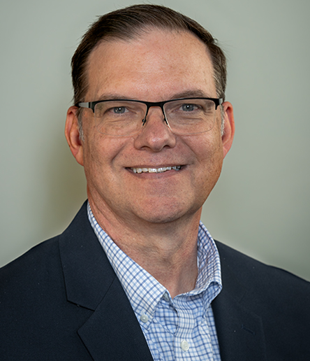Chris Crifasi, a man in glasses, dons a dark blazer and checkered shirt while smiling warmly at the camera against a plain background.