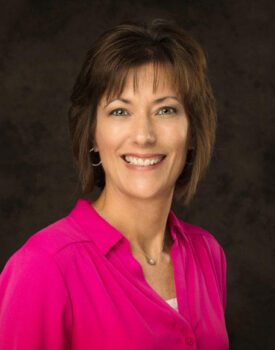 Dana Steffens, a woman with short brown hair and hoop earrings, radiates joy in her bright pink blouse as she smiles against a dark background.