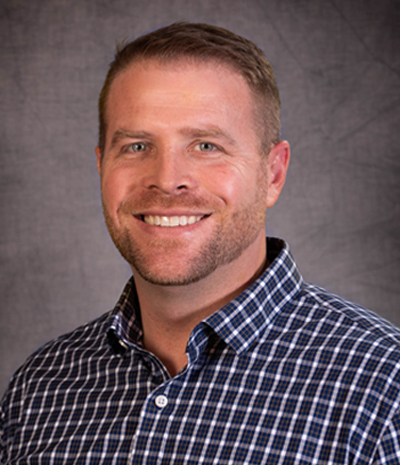 A man with short hair and a beard, possibly Darin VanVactor, is smiling warmly. He wears a checkered shirt and stands against a gray background.