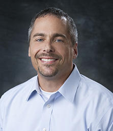 Greg Davis, a man with short hair and a beard, smiles warmly in his light blue shirt against a dark background.