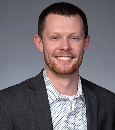 The image features Derek Wagoner, a smiling man in a dark suit and white patterned shirt, standing against a gray background.