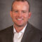 Derrick Hermesch, sporting a light-colored shirt and a brown blazer, smiles warmly against a gray background.