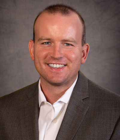Derrick Hermesch, sporting a light-colored shirt and a brown blazer, smiles warmly against a gray background.