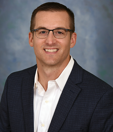 Derrick Deardorff, wearing glasses and a dark blazer over a white shirt, smiles warmly against a neutral, blue-gray background.