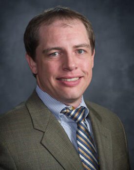 Dusty Marquardt, dressed in a sharp suit and tie, smiles warmly against a gray background.