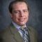 Dusty Marquardt, dressed in a sharp suit and tie, smiles warmly against a gray background.