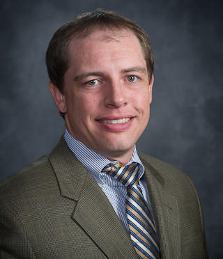 Dusty Marquardt, dressed in a sharp suit and tie, smiles warmly against a gray background.