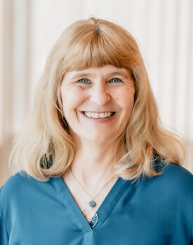 Tamara Egeland, with a radiant smile and shoulder-length blonde hair, stands gracefully in front of a light-colored curtain. Her blue top and necklace complement her vibrant demeanor effortlessly.