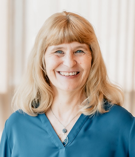 Tamara Egeland, with a radiant smile and shoulder-length blonde hair, stands gracefully in front of a light-colored curtain. Her blue top and necklace complement her vibrant demeanor effortlessly.