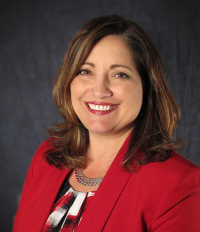 Geri Arndt, a woman with shoulder-length hair in a red blazer and patterned blouse, smiles warmly against a dark background.