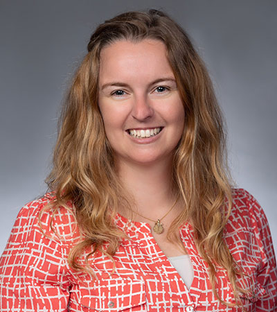 A woman with long wavy hair smiling, wearing a red and white patterned blouse against a gray background.