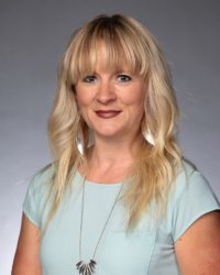 Jessica Stimmel, with long blonde hair, wearing a light blue top and a necklace, poses elegantly against a gray background.
