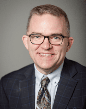 A smiling Marc Johnson, donning glasses, a checkered suit jacket, and a patterned tie, stands against a plain background.