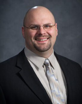 A smiling man, possibly Justin Mentele, dressed in a suit, tie, and glasses is pictured against a dark gray background.