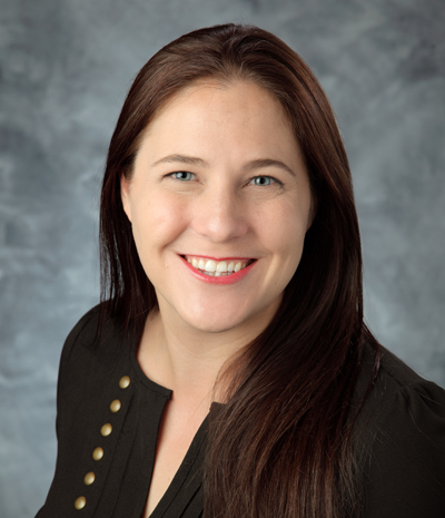Karli Olsen, a smiling individual with long brown hair, is dressed in a dark top set against a gray background.