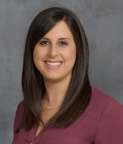 Katherine Laubinger, a smiling woman with straight, dark hair, is elegantly posed in a maroon top and necklace against a gray backdrop.