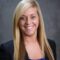 Kati Barnhill, a woman with long blonde hair, is smiling as she dons a black blazer over a blue top against a gray background.