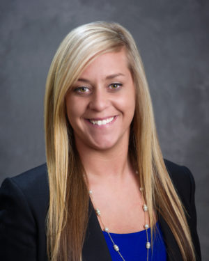 Kati Barnhill, a woman with long blonde hair, is smiling as she dons a black blazer over a blue top against a gray background.