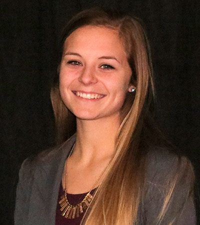 Kaylie Miller, a smiling person with long hair, stands confidently against a dark background, wearing a blazer and necklace.