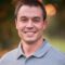 Keaton Dugan, with short dark hair and a gray polo shirt, smiles warmly at the camera against a blurred natural background.